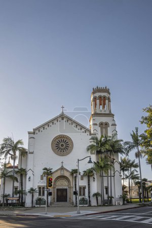Foto de Santa Barbara, CA, EE.UU. - 19 de enero de 2024: Iglesia de Nuestra Señora de los Dolores. Arco esculpido alrededor de madera marrón, engastado en pared blanca con rosas en esquina de calle con torre contra cielo azul - Imagen libre de derechos