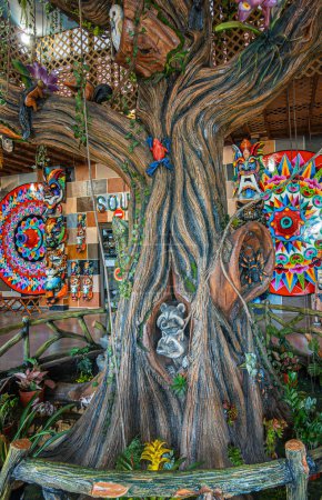 Foto de Costa Rica, San Jerónimo - 22 de julio, 20.23: Tienda de souvenirs El Jardín. Primer plano de la estatua del árbol de madera decorada con imágenes de animales y aves, pared de fondo con mandala colorido y tótem - Imagen libre de derechos