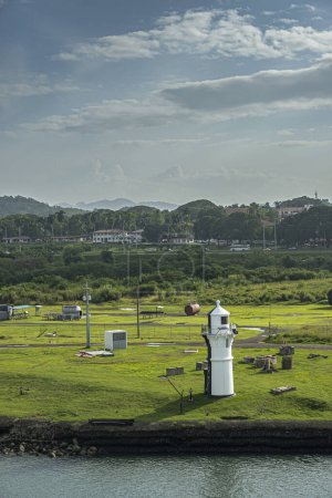 Photo for Panama Canal, Panama - July 24, 2023: White short light beacon on shoreline, set on green lawn, near Miraflores locks under blue cloudscape. Buildings, road with cars and forests in back - Royalty Free Image