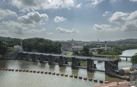 Foto de Canal de Panamá, Panamá - 24 de julio de 2023: Planta termoeléctrica con altas chimeneas en el puente hacia Miraflores bajo un paisaje azul nublado. Planta de energía hidráulica, hidroeléctrica, en frente - Imagen libre de derechos