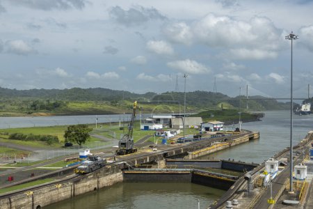Foto de Canal de Panamá, Panamá - 24 de julio de 2023: Las puertas de salida en Pedro Miguel se cierran bajo un paisaje nublado azul. Verdes costas boscosas, buque portacontenedores en el horizonte - Imagen libre de derechos