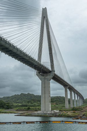 Panama Canal, Panama - July 24, 2023: West giant pylon of Atlantic suspension bridge just out of Canal under gray cloudscape. Colored pipeline floaters in water. Green forested hills