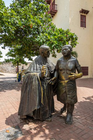 Cartagena, Kolumbien - 25. Juli 2023: Porträt, Statue von San Pedro Claver im Gespräch mit einem Einheimischen auf seinem Platz. Beige historische Fassade und grünes Laub im Rücken.