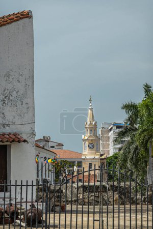 Cartagena, Colombie - 25 juillet 2023 : Tour de l'horloge de la porte principale de la ville vue du bastion de Baluarte de San Ignacio, située entre d'autres grands bâtiments avec des drapeaux et un feuillage vert devant