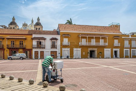Téléchargez les photos : Cartagena, Colombie - 25 juillet 2023 : Vendeur de rue vend des boissons froides sur la Plaza de la Aduana. Santuario de San Pedro Claver top avec des tours et des coups d'oeil horloge sur les maisons de 2 étages sous le ciel bleu - en image libre de droit