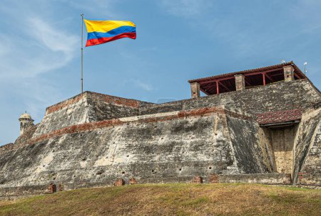 Cartagena, Kolumbien - 25. Juli 2023: Nahaufnahme, grau-beiger Stein San Felipe de Barajas Festungsmauern und Nationalflagge. Gegen blaue Wolken.