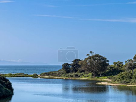 Photo for Santa Barbara, California, USA - March 17, 2024: UCSB, Sandy beach dam, with dune vegetation separates Campus Lagoon from Pacific Ocean under sunny blue sky. Green foliage on sides - Royalty Free Image