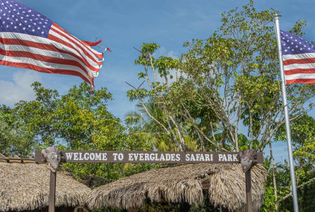 Everglades, Florida, USA - July 29, 2023: Safari park welcome sign with national flag above and backed by green foliage under blue sky. Fake aligator heads as dedoration