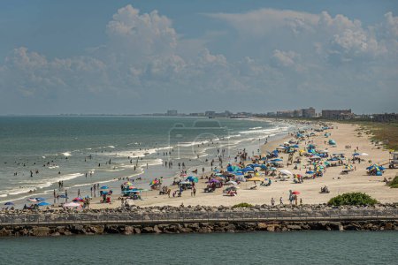 Port Canaveral, Florida, USA - July 30, 2023: Long sandy beach, full of bathers in water and on sand, stretches out as far as one can see, south  of Fisherman Pier under blue cloudscape