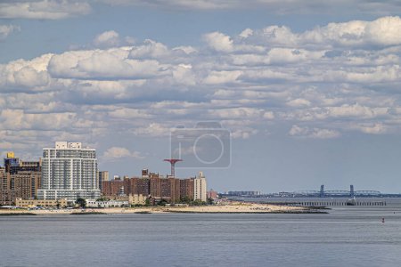 New York, NY, USA - 1. August 2023: Der Strand von Coney Island mit der Gil Hodges Memorial Bridge im Hintergrund unter blauer Wolkendecke. Maimonides Park-Turm ragt über hohe Hauswand