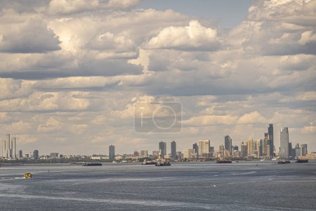 New York, NY, USA - August 1, 2023: New Jersey and Manhattan skyline with Miss Liberty statue in middle  seen from The Narrows bay under dense blue cloudscape. Vessels on the water