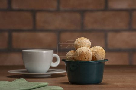 Foto de Cheese breads in a green ramekin with a cup of coffee on wooden table and bricks wall background. - Imagen libre de derechos