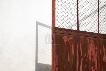 A brown metal gate with peeling paint falling a shadow on the white wall. Negative space