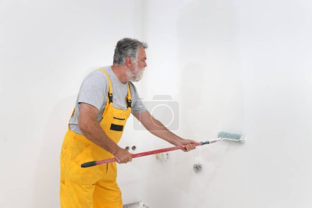 Foto de Senior adulto caucásico trabajador pintura pared a blanco con rodillo de pintura - Imagen libre de derechos