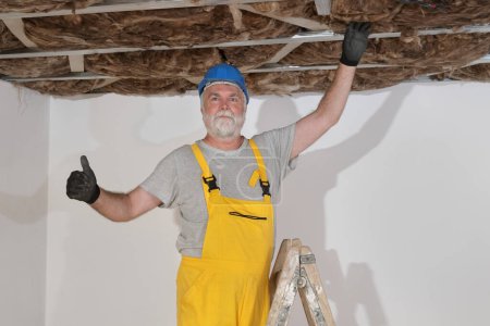 Foto de Trabajador adulto mayor que coloca aislamiento de lana mineral para la eficiencia energética al techo con la construcción de aluminio y el gesto con el pulgar hacia arriba - Imagen libre de derechos