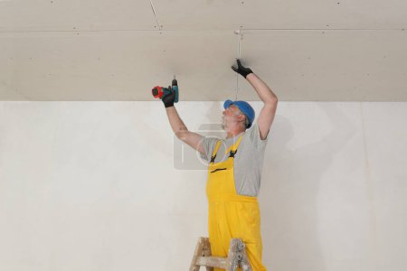 Foto de Trabajador fijación de techo de yeso drywall utilizando destornillador de peaje de energía eléctrica, placas de yeso sobre aislamiento de lana mineral para la eficiencia energética - Imagen libre de derechos