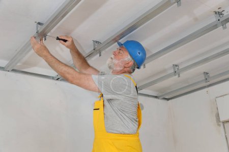 Foto de Trabajador adulto mayor colocando perfil de aluminio para techo de yeso y aislamiento usando destornillador - Imagen libre de derechos