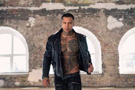 Foto de Hot tattooed man in leather against the big window.Handsome Young Athletic Male Fashion Model.Muscular athletic sexy male with naked torso. Brutal handsome man with tattooed body. - Imagen libre de derechos
