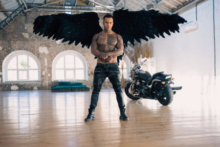 Foto de Angel man with black wings, stand posing at camera. man fall from heaven, angel with muscular body gained freedom.  tattoed hot man. tattooed black angel. dark angel in leather - Imagen libre de derechos