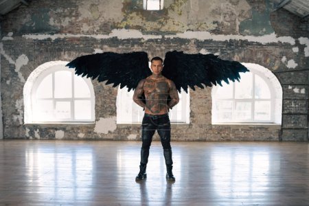 Foto de Angel man with black wings, stand posing at camera. man fall from heaven, angel with muscular body gained freedom.  tattooed hot man. tattooed black angel. dark angel in leather - Imagen libre de derechos