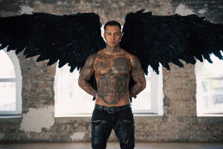 Photo for Angel man with black wings, stand posing at camera. man fall from heaven, angel with muscular body gained freedom.  tattooed hot man. tattooed black angel. dark angel in leather - Royalty Free Image