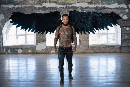 Téléchargez les photos : Angel man with black wings, stand posing at camera. man fall from heaven, angel with muscular body gained freedom.  tattooed hot man. tattooed black angel. dark angel in leather - en image libre de droit
