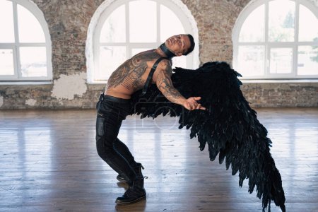 Foto de Handsome man with wings.Male angel with black wings.  Muscular shirtless man with whip. Brutal handsome man with tattooed body. Muscular athletic sexy male - Imagen libre de derechos