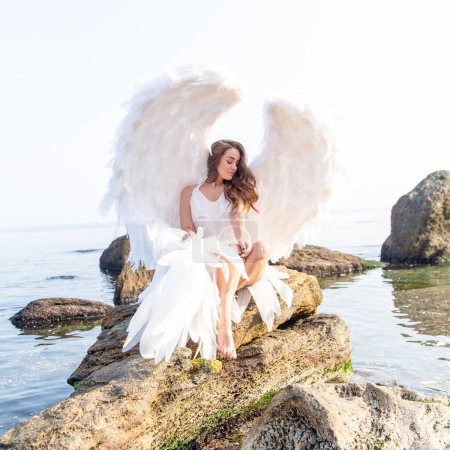 Foto de Hermoso ángel con grandes alas blancas. Mujer tierna joven en vestido de seda blanca caminando en la orilla del mar - Imagen libre de derechos