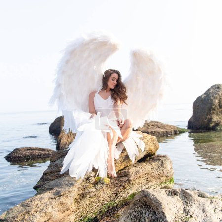 Foto de Hermoso ángel con grandes alas blancas. Mujer tierna joven en vestido de seda blanca caminando en la orilla del mar - Imagen libre de derechos