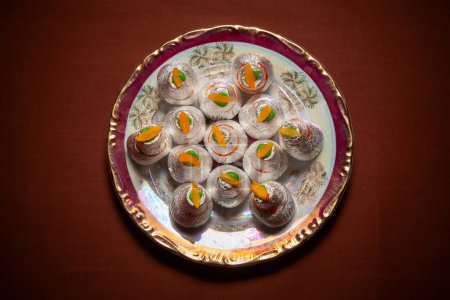 Top view of an Indian Traditional sweet called "khoya Mithai" an Indian Sweet garnished with silver leaves and dry fruits." served on a ceramic plate on a dark brown background.