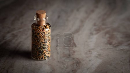 A small glass bottle filled with a blend of five spices called "Pachphoran" is placed on a marble background.