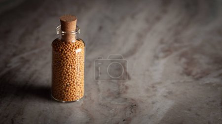A small glass bottle filled with organic Mustard seed (Sinapis alba) is placed on a marble background.