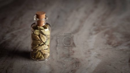 Eine kleine Glasflasche mit organischem Kardamom oder Kardamom (Elettaria cardamomum) wird auf einem Marmorgrund platziert.