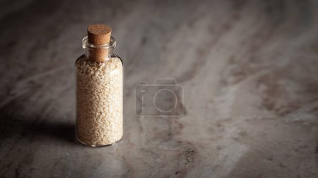 Téléchargez les photos : Une petite bouteille en verre remplie de graines de sésame blanc bio (Sesamum indicum) ou de Til est placée sur un fond de marbre.. - en image libre de droit