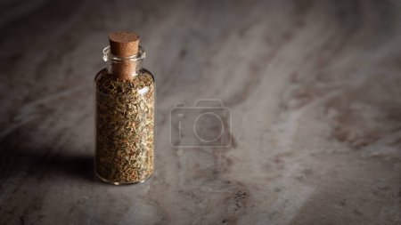 A small glass bottle filled with dried "Oregano leaves" placed on a marble background.