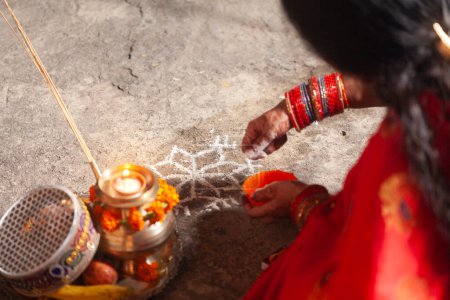 An Indian woman is making an Auspicious Hindu design pattern (rangoli) for Karwa Chauth in a red saree.