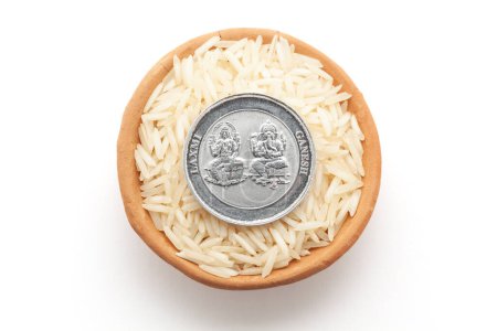 Blick von oben auf einen irdenen Topf, gefüllt mit Reis (Oryza sativa). Eine Silbermünze mit eingravierten Hindu-Gottheiten Ganesha und Laxmi. Isoliert auf weißem Hintergrund.