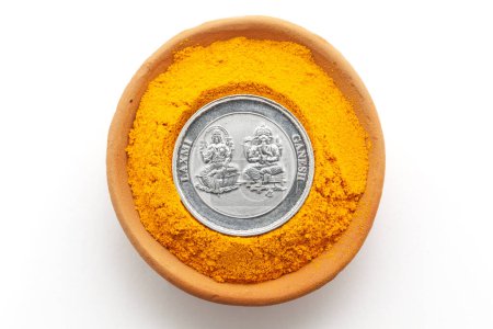 Blick von oben auf einen irdenen Topf, gefüllt mit Kurkuma (Haldi). Eine Silbermünze mit eingravierten Hindu-Gottheiten Ganesha und Laxmi. Isoliert auf weißem Hintergrund.