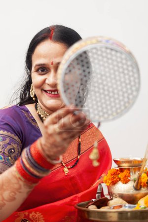 Foto de Una mujer india alegre celebra Karwa Chauth, un festival hindú, vistiendo un saree tradicional y colorido y mirando a través de un tamiz. - Imagen libre de derechos