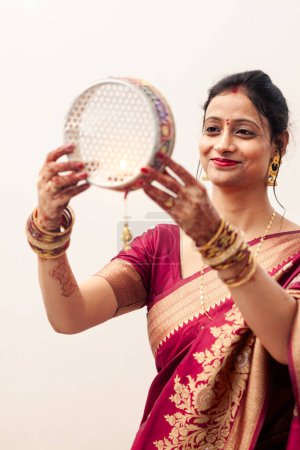 Téléchargez les photos : Une Indienne joyeuse célèbre Karwa Chauth, une fête hindoue, portant un saree traditionnel et coloré et regardant à travers un tamis. - en image libre de droit