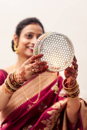 Téléchargez les photos : Une Indienne joyeuse célèbre Karwa Chauth, une fête hindoue, portant un saree traditionnel et coloré et regardant à travers un tamis. - en image libre de droit