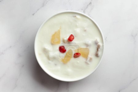 Close-up of Fruits raita or Yogurt & Pineapple or Ananas Raita garnished with pineapple piece and pomegranate, served in a white ceramic bowl over whiet granite background