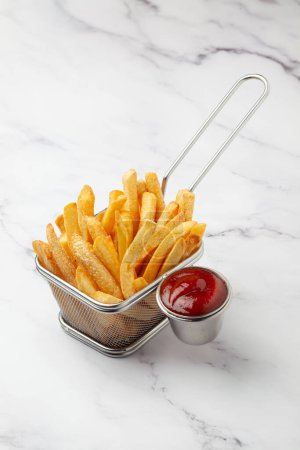 Primer plano de papas doradas papas fritas crujientes servidas en la canasta con salsa de tomate rojo sobre fondo de granito blanco.