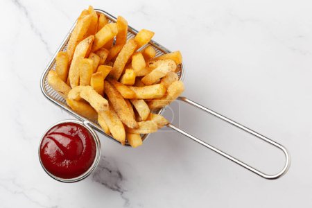 Primer plano de papas doradas papas fritas crujientes servidas en la canasta con salsa de tomate rojo sobre fondo de granito blanco.