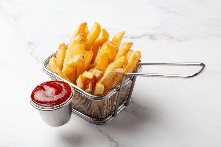 Nahaufnahme von goldenen Kartoffeln Crispy Pommes im Korb serviert mit rotem Tomatenketchup über weißem Granithintergrund.