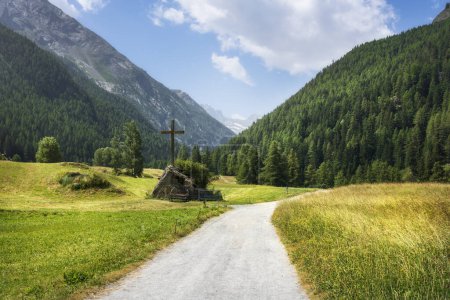 Chemin de montagne et croix chrétienne à Prati di Sant'Orso. Massif Gran Paradiso en arrière-plan. Cogne, Vallée d'Aoste, Italie