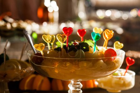 Foto de Frutas recién cortadas, dispuestas en un jarrón de vidrio con palillos de plástico en forma de corazón. Los frutos incluyen manzanas, naranjas, plátanos y uvas. - Imagen libre de derechos