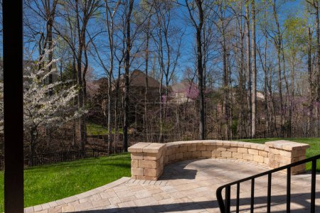 Backyard view in spring season with patio pavers and stone wall, blooming white cherry tree, and spring colored woods in the background.