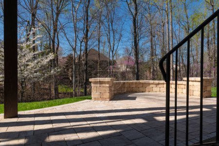 Photo for Picturesque backyard view in spring season with patio pavers and stone wall, blooming white cherry tree, and spring colored woods in the background. - Royalty Free Image