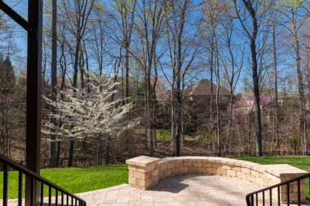 Pintoresca vista del patio trasero en temporada de primavera con adoquines de patio y pared de piedra, cerezo blanco en flor, y maderas de color primavera en el fondo.
